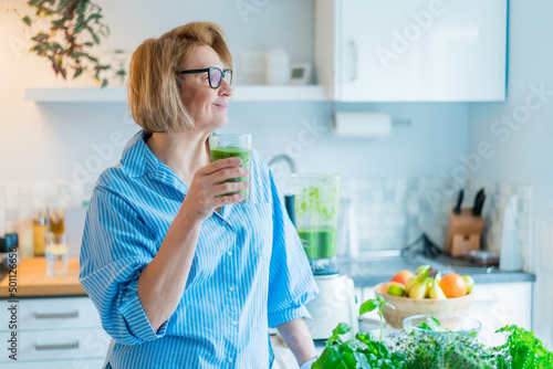 Smiling middle aged woman with just made glass of detox shake, green smoothie in the kitchen. Healthy dieting, eating, cooking. Natural anti aging methods, weight loss program. Vegan, vegetarian diet photo