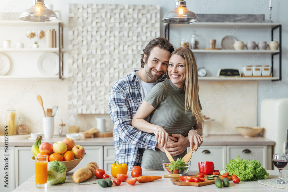 Cheerful young caucasian lady cooking salad, guy with stubble hugs woman at table with organic vegetables Cheerful young caucasian lady cooking salad, guy with stubble hugs woman at table with organic vegetables