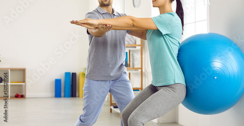 Young female patient together with physiotherapist does exercises for back with help of fitball. Cropped image woman perform squats with fitball to get rid of back pain and restore spine health.