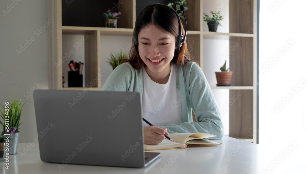 Happy young woman in headphones speaking looking at laptop making notes, girl student talking by video conference call, female teacher trainer tutoring by webcam, online training, e-coaching concept Happy young woman in headphones speaking looking at laptop making notes, girl student talking by video conference call, female teacher trainer tutoring by webcam, online training, e-coaching concept