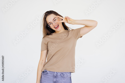 Young woman having fun, showing v-sign over eye and winking, smiling sassy at camera, being in good and positive mood, standing over white background