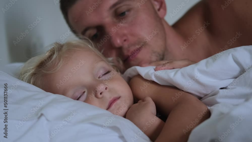 The father kisses his sleeping daughter on the shoulder, gently looking at her, then kisses her on the head again, covers his daughter with a blanket and calmly falls asleep next to her in bed. The father kisses his sleeping daughter on the shoulder, gently looking at her, then kisses her on the head again, covers his daughter with a blanket and calmly falls asleep next to her in bed.