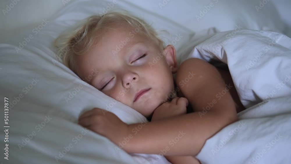 A blonde girl of two years old sleeps soundly, peacefully in a warm bed on a snow-white pillow, covered with a snow-white blanket. A little girl has pleasant dreams. Healthy baby sleep. A blonde girl of two years old sleeps soundly, peacefully in a warm bed on a snow-white pillow, covered with a snow-white blanket. A little girl has pleasant dreams. Healthy baby sleep.
