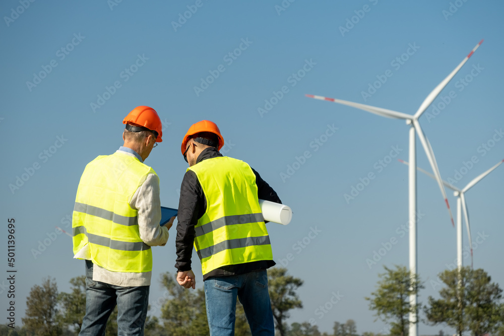 Propellers rotate against cloudless sky and trees. Professional technicians of maintenance control operation of wind farm with tablet back view