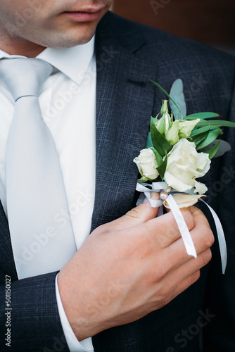 A stylish groom in a blue suit, tie and white shirt touches a boutonniere of white roses and leaves.