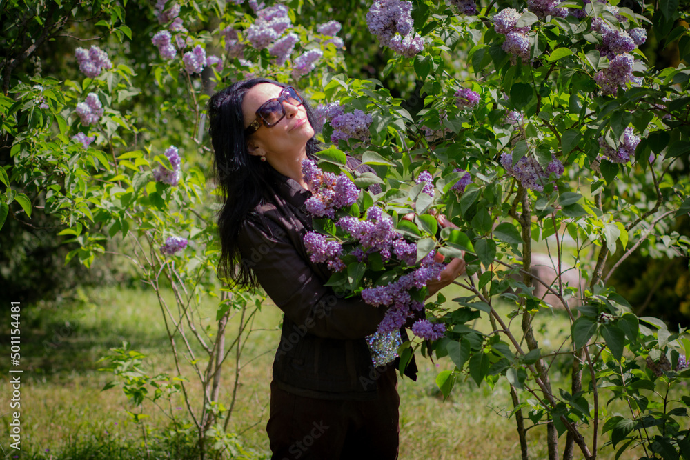 A happy girl with long black hair in glasses hugs and sniffs lilac branches in the garden. Rest in the park. A happy girl with long black hair in glasses hugs and sniffs lilac branches in the garden. Rest in the park.