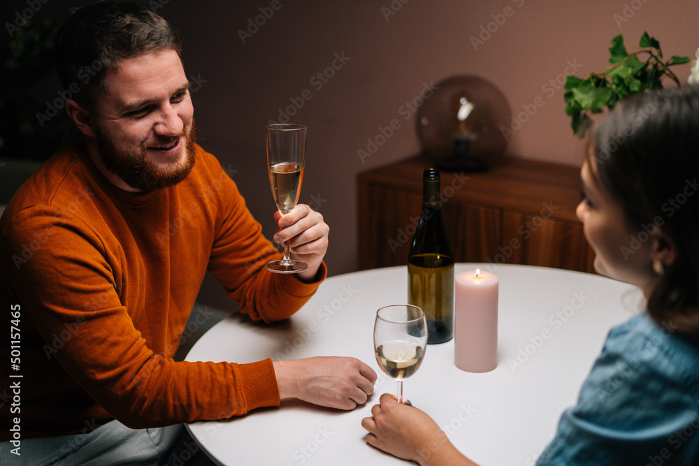 High-angle view of smiling bearded young man talking celebration toast and clinking white wine glass with girlfriend during romantic dinner. Love couple celebrating anniversary or Valentines day. High-angle view of smiling bearded young man talking celebration toast and clinking white wine glass with girlfriend during romantic dinner. Love couple celebrating anniversary or Valentines day.