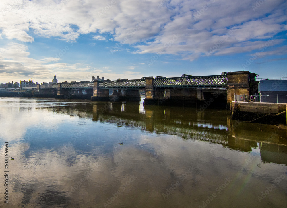 The Rochester Railway Bridge over the River Medway, across the water from the North East bank.  A cold blue sky and a few clouds.  The reflection of the bridge is seen in the river.