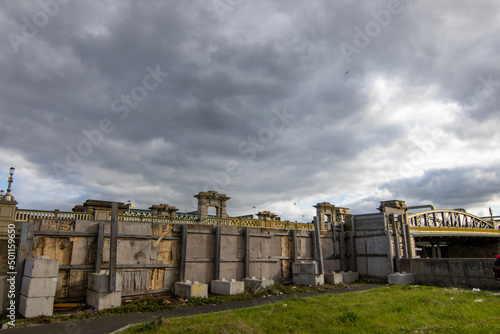 Beside the Rochester Bridge over the River Medway, are these wooden hoardings, with concrete blocks. The stone architecture and railings of the Edwardian bridge can be seen below storm clouds building
