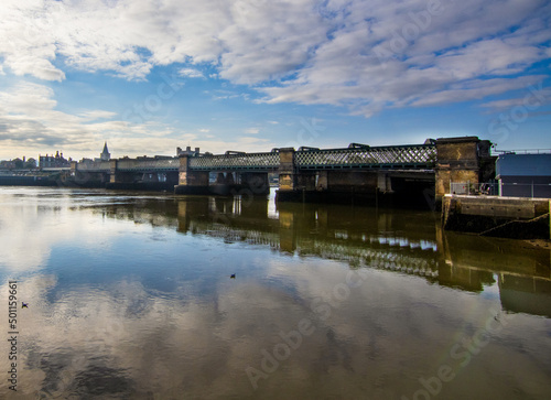 The Rochester Railway Bridge over the River Medway, across the water from the North East bank.  A cold blue sky and a few clouds.  The reflection of the bridge is seen in the river.