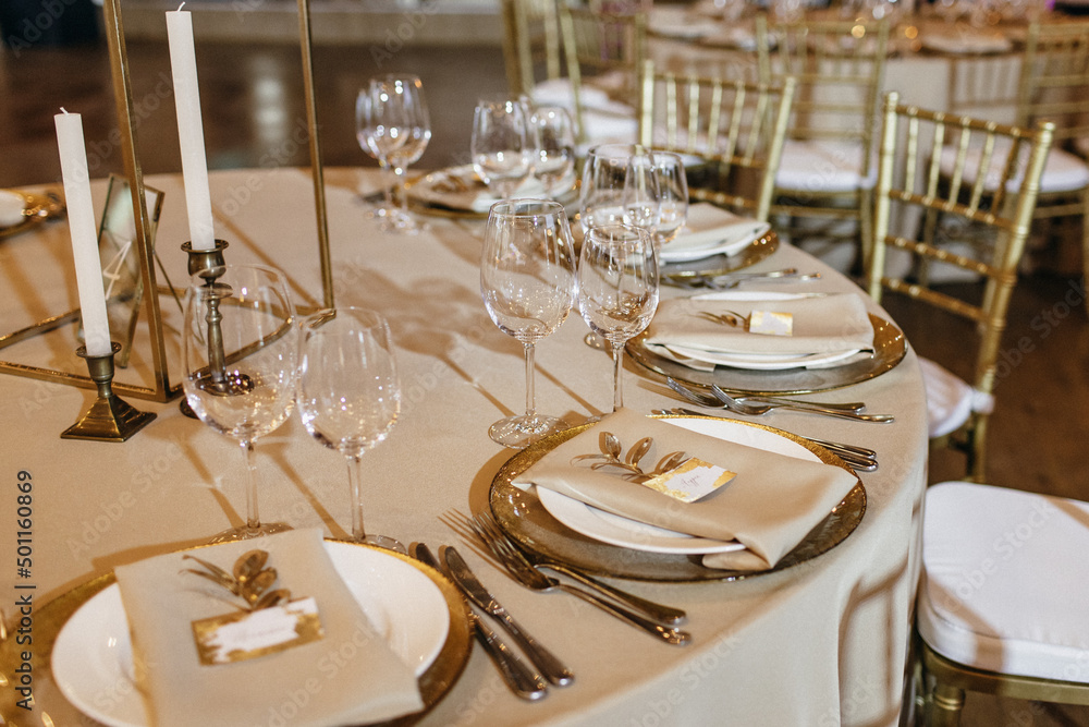 On the table in wedding banquet area are gold plates, napkins, cutlery, glasses, there are candles, menu cards lie on the plates.