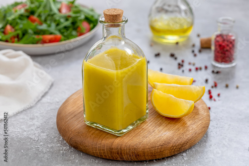 Salad dressing with oil, lemon juice, salt and pepper served in glass cruet or bottle. Close up, ingredients and plate with arugula and tomato salad on background. Horizontal. Basic Vinaigrette. photo