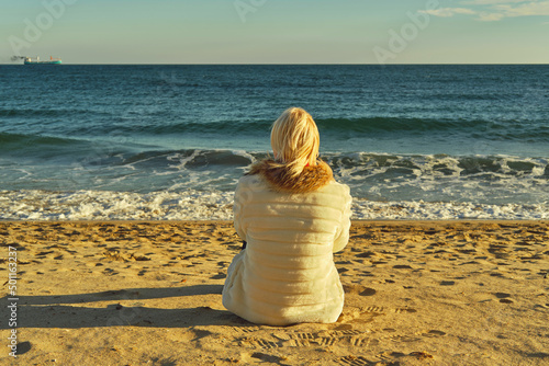 A middle-aged woman with blonde hair sits on a sandy beach looking out to sea. 