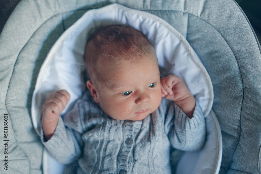 Cute European baby in knitted suit, 1 month old baby lies in swing, top view. Baby 2 months old with open eyes looking at camera Cute European baby in knitted suit, 1 month old baby lies in swing, top view. Baby 2 months old with open eyes looking at camera