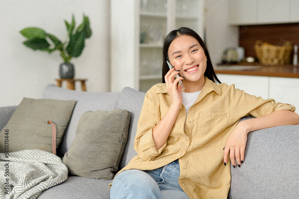 Smiling young woman chatting with friend on mobile phone, has pleasure phone conversation. Charming asian woman talking on the smartphone sitting on the cozy couch at home Smiling young woman chatting with friend on mobile phone, has pleasure phone conversation. Charming asian woman talking on the smartphone sitting on the cozy couch at home