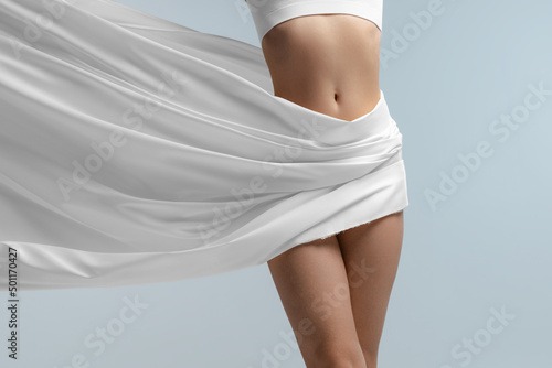 Print op canvas Cropped portrait of slim, smooth female body in white lingerie with silk fabrik isolated over blue studio background