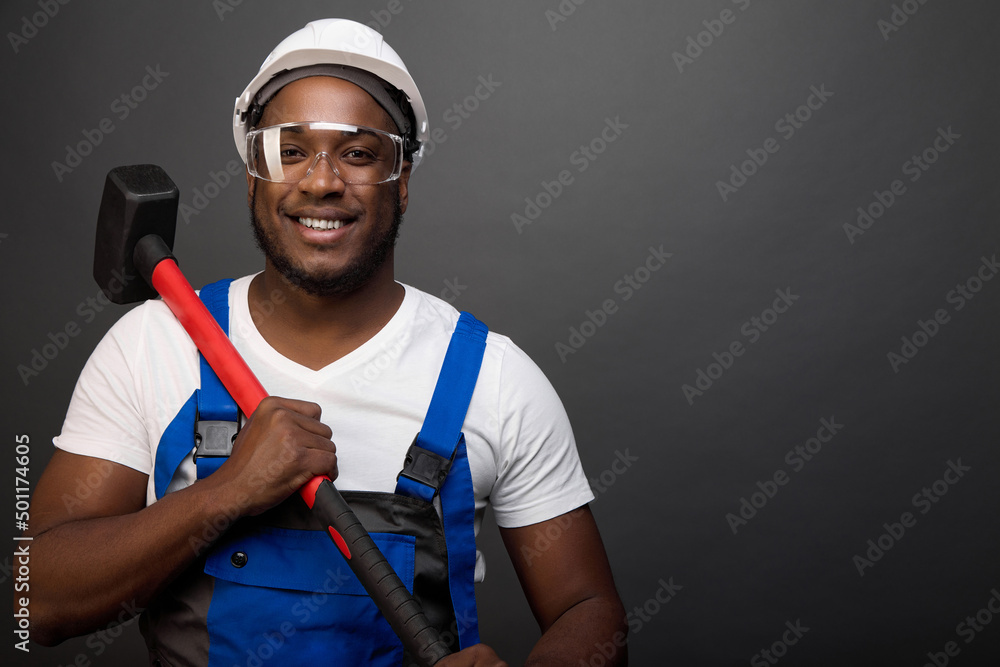 Strong black worker holds a heavy sledgehammer with a red handle. An iron hammer is carried on a strong shoulder by a courageous African-American construction worker in protective glasses and a helmet Strong black worker holds a heavy sledgehammer with a red handle. An iron hammer is carried on a strong shoulder by a courageous African-American construction worker in protective glasses and a helmet