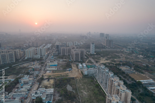 aerial drone shot passing over a building with homes, offices, shopping centers moving towards skyscapers in front of sunset showing the empty outskirts of the city of gurgaon