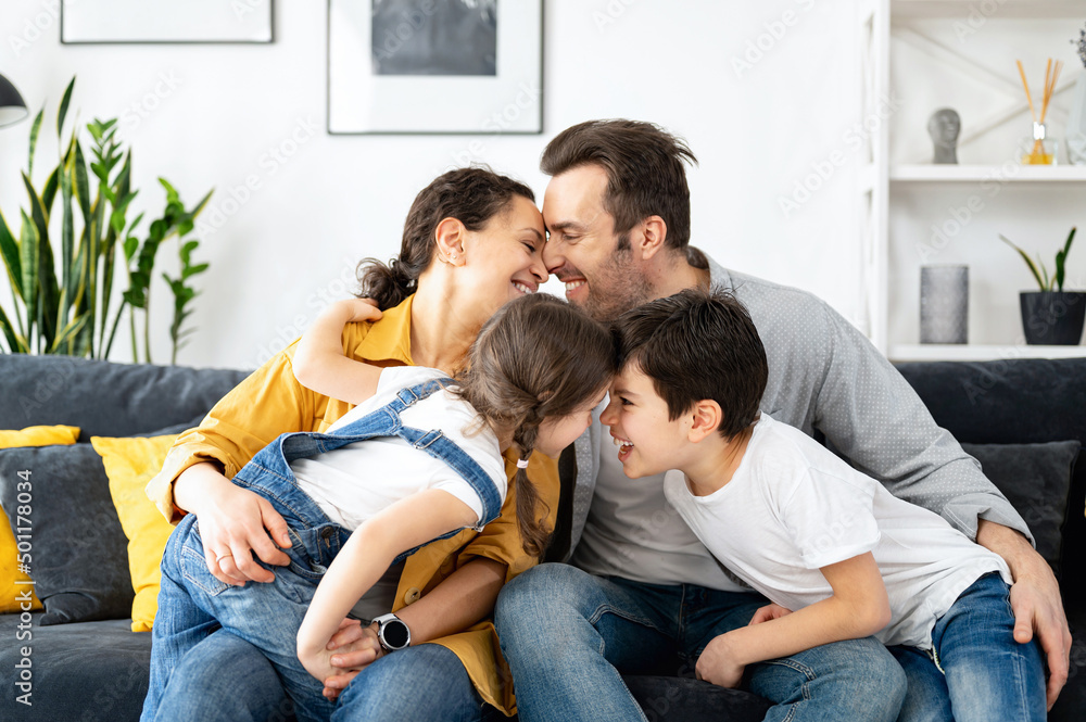 Cheerful multiracial family of four - mom, dad, son and toddler daughter sitting at the sofa in cozy living room, bonding to each other, enjoying weekend at home