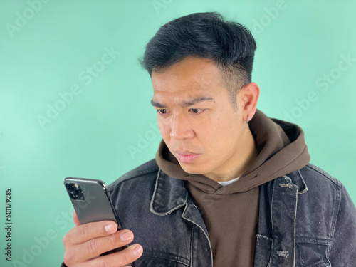 Handsome young Asian man using his mobile phone with a surprised face, wearing casual clothes and green background. Active youth concept. © dianix!
