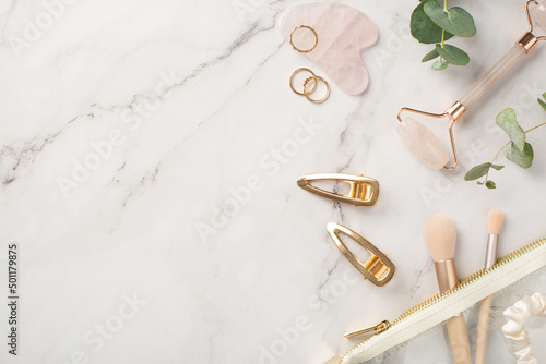 Top view photo of golden barrettes rose quartz roller gua sha golden rings makeup brushes scrunchy transparent cosmetics bag and eucalyptus on white marble background with empty space