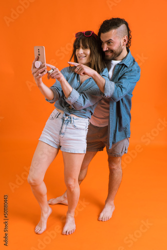 Two excited people using mobile phones while standing at the studio with orange background and pointing finger