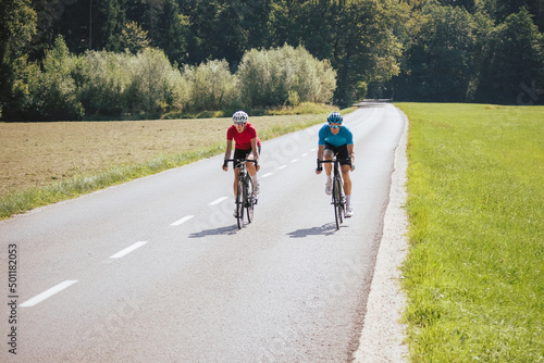 Fototapeta Naklejka Na Ścianę i Meble -  Front view of a woman and man, professional racing bikers in sportswear, riding along an asphalt road in the beautiful green nature.