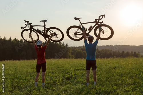 Two cyclers silhouettes holding road bicycles high above heads, in winner pose during sunset over the mountain landscape
