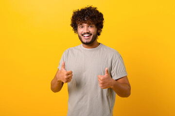 Like, excellent job. Pleased Indian man smiling to camera and gesturing thumbs up, approval hand sign, satisfied with result. Indoor studio shot isolated on yellow background