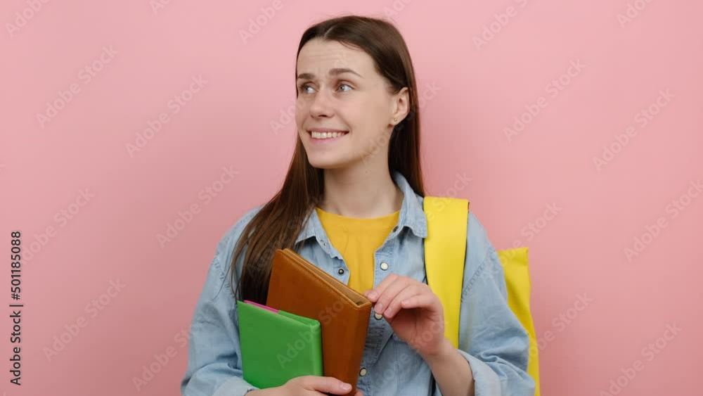 Portrait of confused fun girl teen student hold books spreading hands say oops ouch oh omg i am so sorry, wears shirt and yellow backpack, posing isolated over pink color background wall in studio Portrait of confused fun girl teen student hold books spreading hands say oops ouch oh omg i am so sorry, wears shirt and yellow backpack, posing isolated over pink color background wall in studio