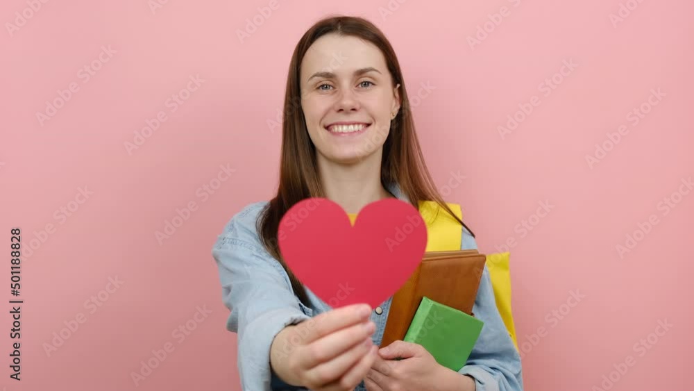 Happy girl teen student wears shirt and yellow backpack hold books showing red heart, smiling looking at camera, isolated on pink color background studio. Education in university college concept Happy girl teen student wears shirt and yellow backpack hold books showing red heart, smiling looking at camera, isolated on pink color background studio. Education in university college concept
