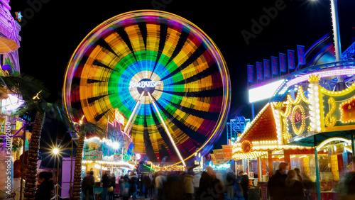 Soest, Germany - November.05.2022: Colorfully illuminated ferris wheel at a funfair at night with abstract motion blur due to long time exposure and food stands in the foreground