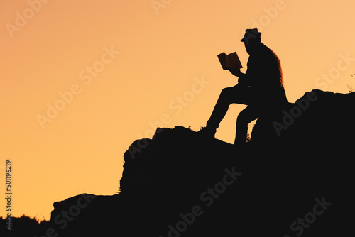 silhouette of a man in a hat reading a book sitting on a mountainside