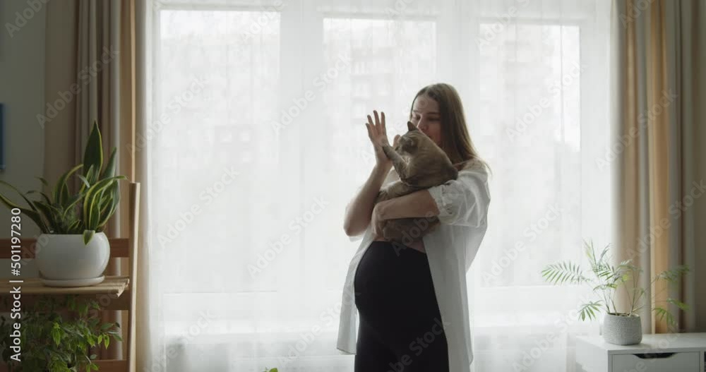 Happy cheerful pregnant woman dances with a cat in her arms near the window at home. Expectant loving mother relax at home listening to the music. Maternity prenatal care and woman pregnancy concept. Happy cheerful pregnant woman dances with a cat in her arms near the window at home. Expectant loving mother relax at home listening to the music. Maternity prenatal care and woman pregnancy concept.
