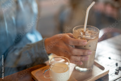 A hand holding a tall glass of iced latte coffee with milk on a wooden bar over a cafe glass window reflex at a Cafe coffee shop. Cold brew refreshment summer drink with copy space. Selective focus