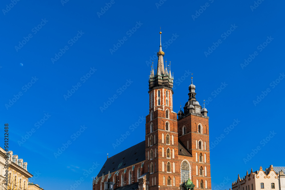 St. Mary's Basilica on the main market square of Krakow on a sunny day, the moon and the trail of a jet plane are visible in the blue sky. Tourist travel to European attractions