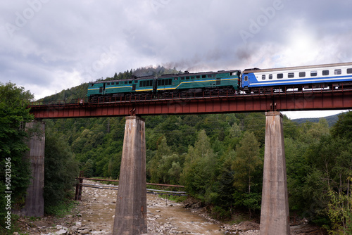 An old diesel train with passenger cars moves along the bridge over the mountain slopes above the seething river. Ukraine. Carpathian mountains. photo