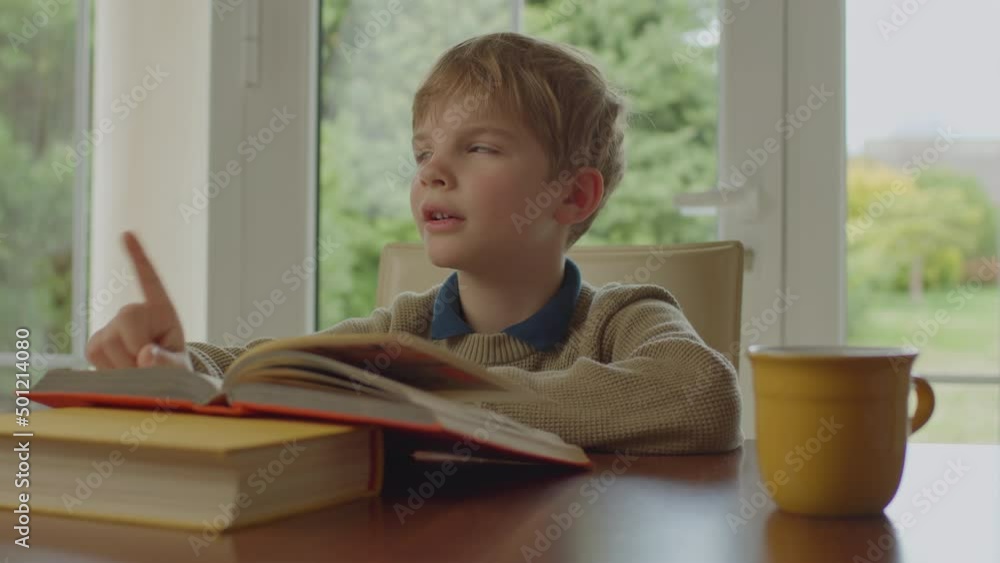 School kid reading paper book sitting alone at the table at home. Cute blonde boy learning at home. School kid reading paper book sitting alone at the table at home. Cute blonde boy learning at home.