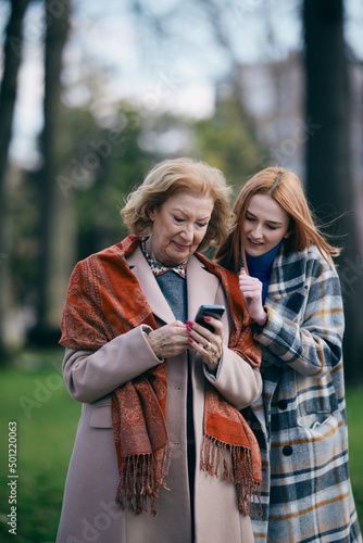 A grandmother is browsing her phone during a walk in the park with her adolescent granddaughter in cold weather.