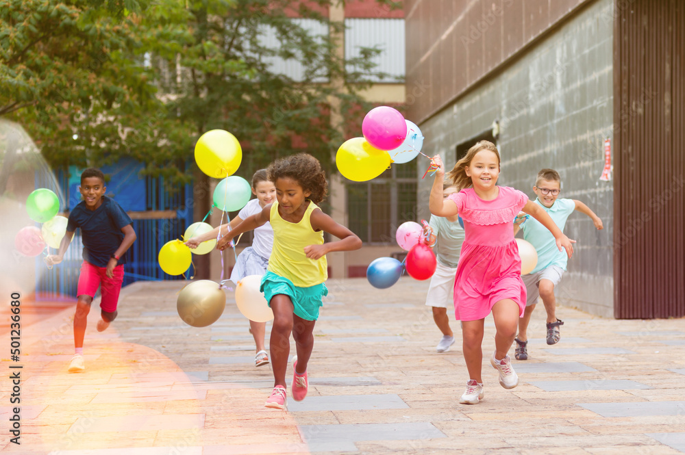 Portrait of cheerful preteen girls and boys with colorful toy balloons in hands running on city street on sunny summer day - obrazy, fototapety, plakaty 