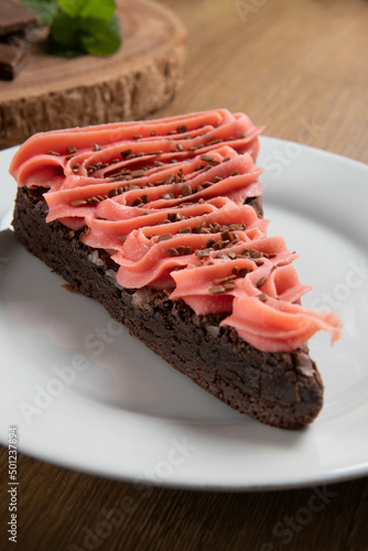Piece of Chocolate Brownie Slice with Strawberry and Chocolate Icing. Wooden table with mint and chocolate chips in the background