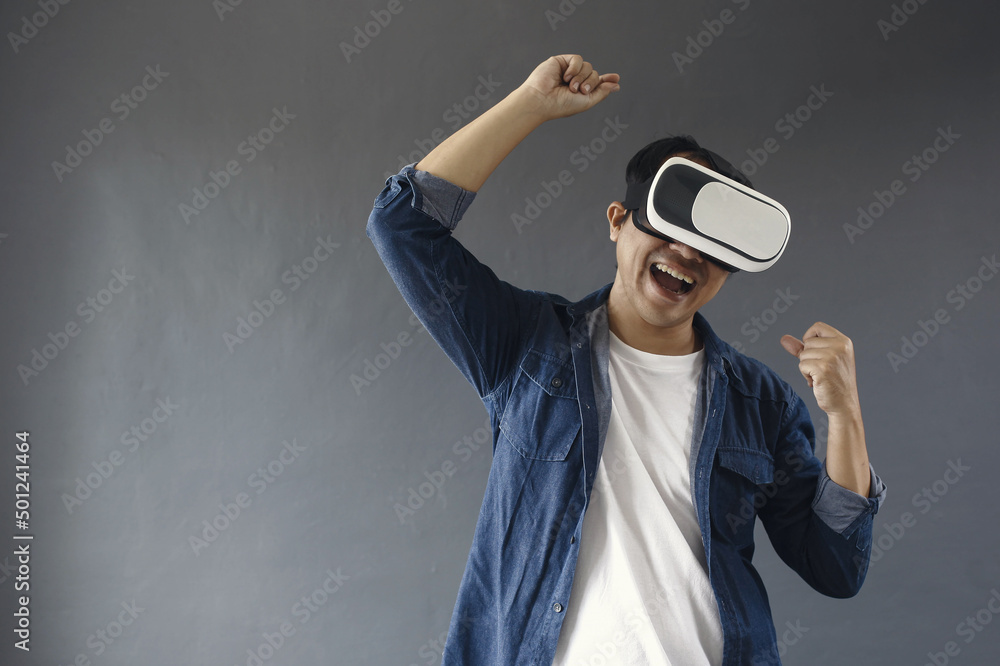 Enthusiastic asian man using VR is celebrating victory, smiling saying yes, winning game or competition, achieve goal, dark grey background Enthusiastic asian man using VR is celebrating victory, smiling saying yes, winning game or competition, achieve goal, dark grey background