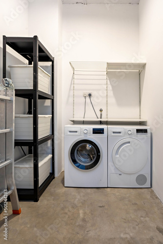 Interior of a storeroom with modern washing machine, dryer machine, foldable ladder, shelf with tools and boxes.