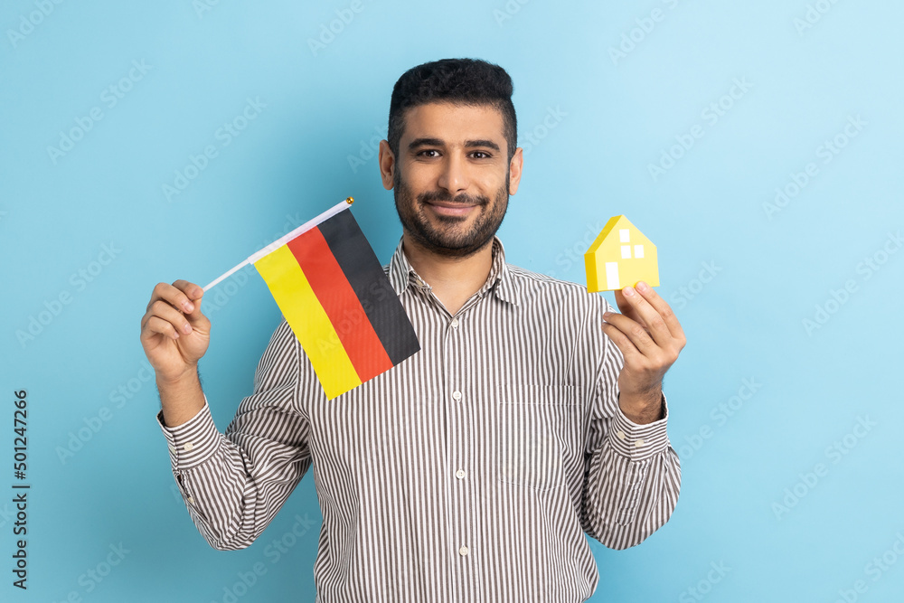 Satisfied man standing with paper house and flag of German, immigration and house purchase, rental housing, wearing striped shirt. Indoor studio shot isolated on blue background. Satisfied man standing with paper house and flag of German, immigration and house purchase, rental housing, wearing striped shirt. Indoor studio shot isolated on blue background.