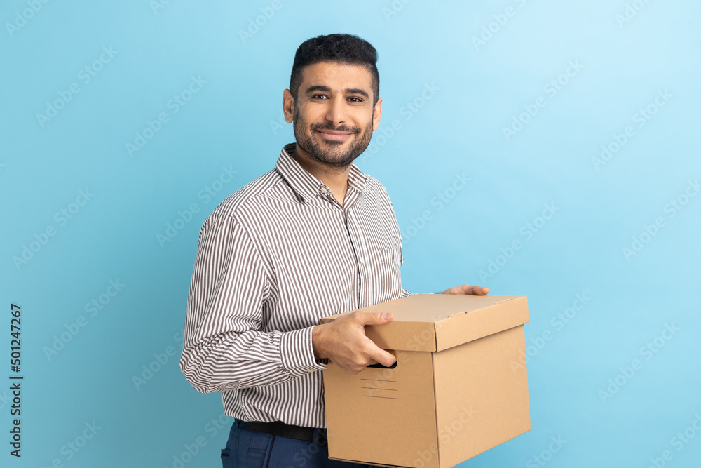Smiling positive businessman holding cardboard parcel, delivering orders to address, looking at camera with friendly expression, wearing striped shirt. Indoor studio shot isolated on blue background. Smiling positive businessman holding cardboard parcel, delivering orders to address, looking at camera with friendly expression, wearing striped shirt. Indoor studio shot isolated on blue background.
