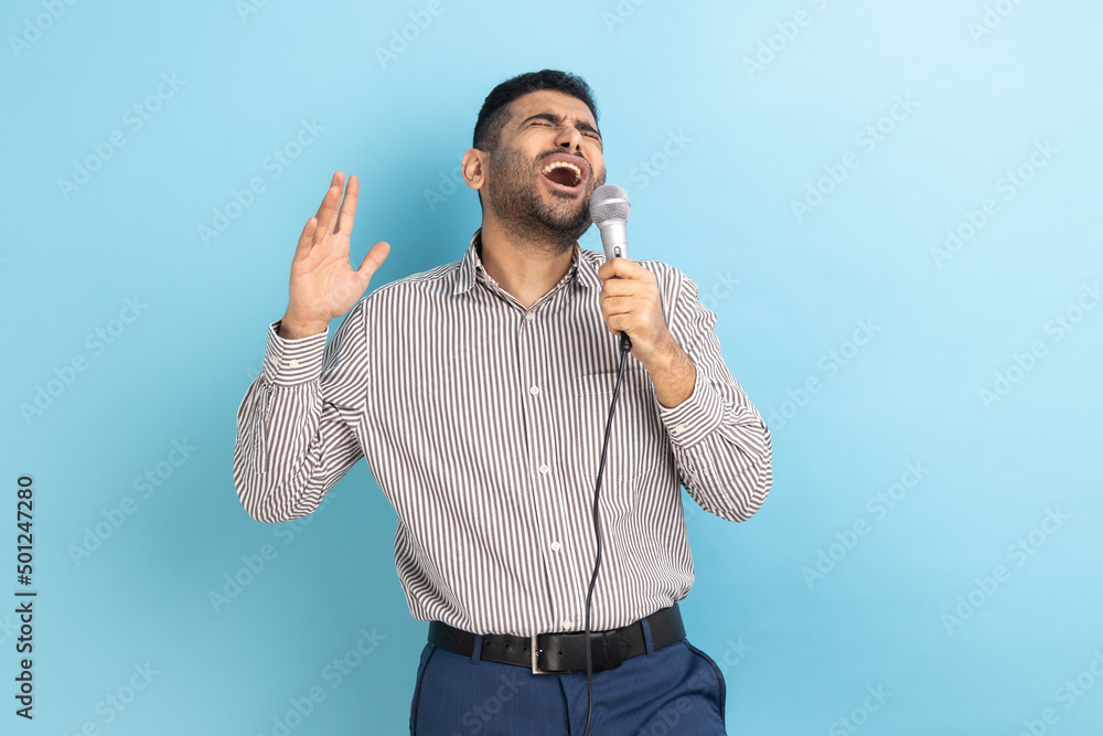 Fototapeta premium Portrait of attractive handsome young adult man with beard singing loud favorite songs with microphone in hands, wearing striped shirt. Indoor studio shot isolated on blue background.
