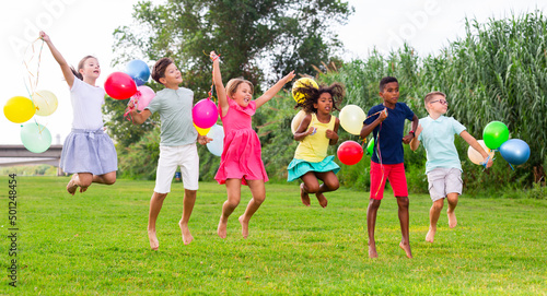 Multiracial group of cheerful emotional preteen friends gaily spending time together on summer day, holding colored helium balloons and jumping together in city park