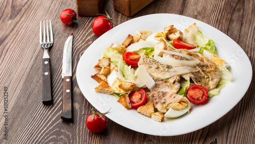 A Caesar salad with grilled chicken breasts, lettuce, croutons, eggs, cherry tomatoes, dressed with Parmesan cheese and sauce. On wooden background.
