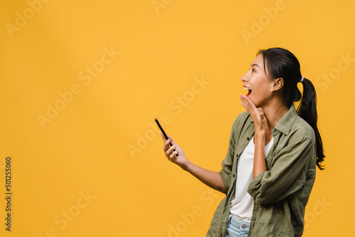 Surprised young Asia lady using mobile phone with positive expression, smiles broadly, dressed in casual clothing and stand isolated on yellow background. Happy adorable glad woman rejoices success.