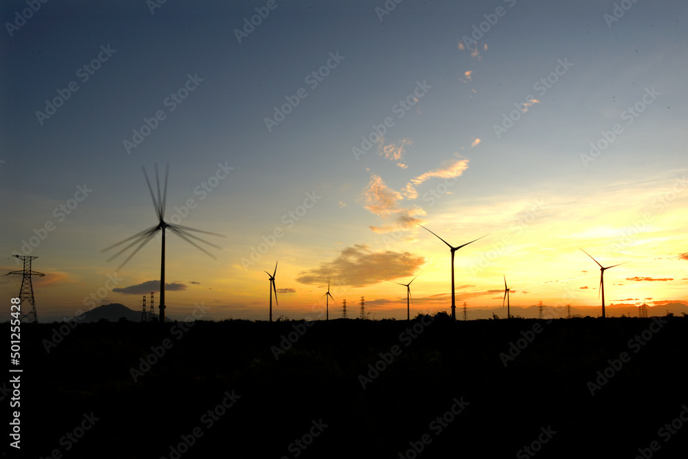 Wind farm or wind park, with high wind turbines in the setting sun for generation electricity with copy space. Green energy concept to reduce climate change and global warming Wind farm or wind park, with high wind turbines in the setting sun for generation electricity with copy space. Green energy concept to reduce climate change and global warming